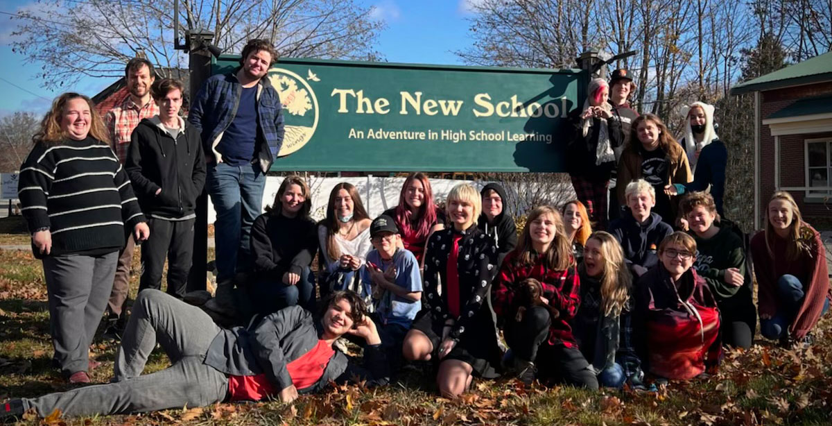 Students and staff pose around the sign in front of The New School. The sign reads "The New School, An Adventure in High School Learning."
