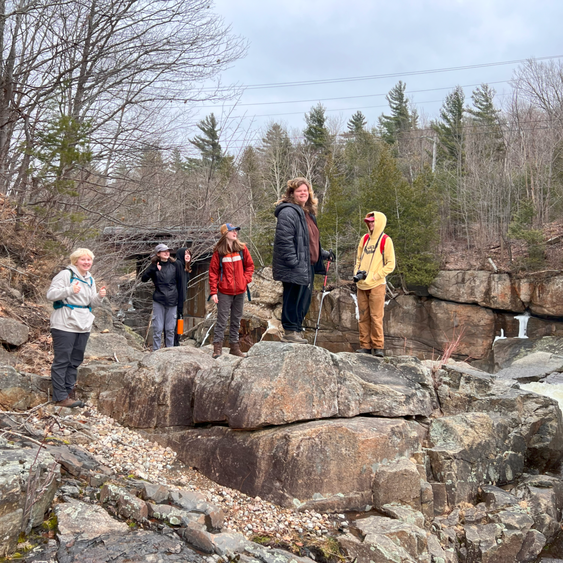 A group of students stand atop boulders by a river.