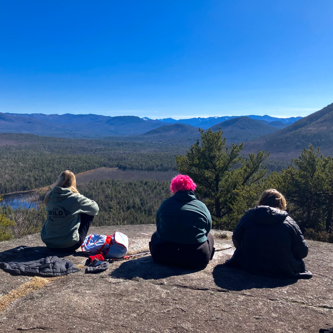 Three students and teachers sit atop a mountain, admiring the view of the valley below under a brilliant blue sky.