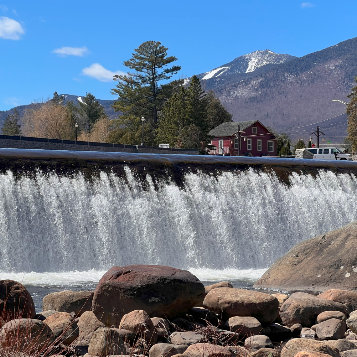A dam with The New School's van parked behind the falls. Above the falls are a red building and several trees. Mountain ridges are in the background.